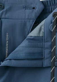 Gros plan de la zone de la fermeture éclair d'une veste bleue avec le texte "MNG Performance", bouton, boutonnière, taille élastique et cordon noir à rayures blanches.