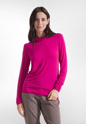 Kvinde med mørkt hår iført en knaldpink top med lange ærmer og taupebukser stående mod en ensartet lys baggrund.