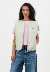 Gilet matelassé vert clair avec fermeture éclair à l'avant et sans manches, porté sur un t-shirt rose, associé à un jean en denim bleu. Détail du logo présent.