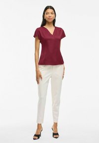 Blusa bordeaux a maniche corte con scollatura a V, abbinata a pantaloni su misura color crema e tacchi a vista neri. Texture liscia, silhouette aderente.