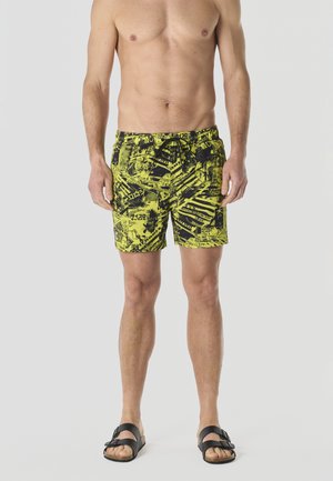 Hombre de pie vistiendo pantalones cortos de baño gráficos en amarillo y negro y sandalias negras con hebilla, sobre un fondo gris claro liso.