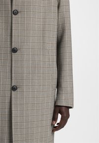Cappotto a quadri in tonalità beige e marrone, realizzato in tessuto testurizzato. Presenta bottoni neri e una tasca laterale, con una vestibilità sartoriale.