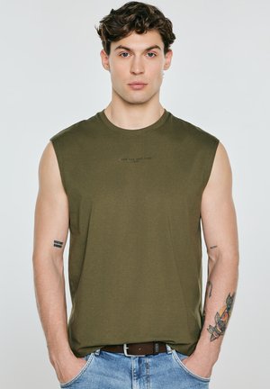 Młody mężczyzna z kręconymi brązowymi włosami nosi bezrękawnik oliwkowej zieleni i niebieskie dżinsy, pokazując tatuaże na obu przedramionach na jednolitym tle.