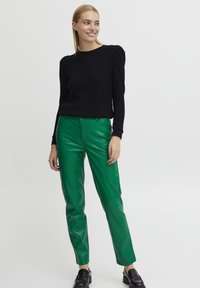  mujer que lleva una blusa negra de mangas largas, pantalones de cuero verde brillante y mocasines negros, de pie contra un fondo blanco liso.