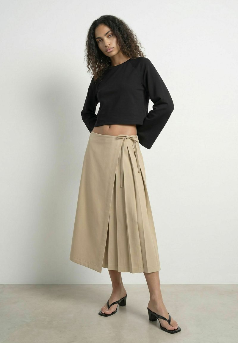 Femme aux cheveux bouclés portant un crop top noir à manches longues, une jupe portefeuille beige et des sandales à talons noires, debout devant un fond uni.