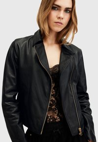 Giacca da motociclista nera in pelle con chiusura a zip, colletto a punta e design aderente. Presenta dettagli in oro e una texture liscia.