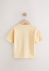 Schlichtes hellgelbes T-Shirt für Kleinkinder mit kurzen Ärmeln, hängend an einem Holzkleiderbügel vor einer weißen Wand, Rückansicht.