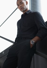 Chemise noire à manches longues, associée à un pantalon noir. Montre en argent visible au poignet, se tenant devant un fond en verre.