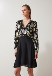 Svart klänning med en blommig guldmönstrad överdel, V-ringning och långa ärmar. Utsvängd kjol med en texturerad yta. Elegant och figurnära design.