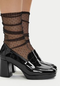 Črni lakasti čevlji z visokimi petami in kvadratnim prstom, s širokim trakcem kot poudarkom. Nošeni s črnimi gležnjčnimi nogavicami iz mrežastega materiala s pikami.