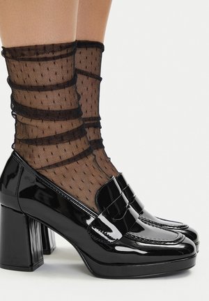 Czarne czółenka na obcasie z lakierowanej skóry, z kwadratowym noskiem i szerokim paskiem jako akcent. Noszone z czarnymi siateczkowymi skarpetkami do kostki w groszki.