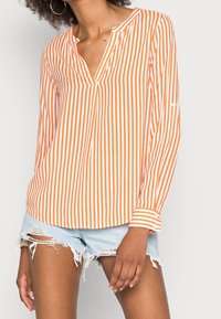 Camicia a maniche lunghe a righe arancioni e bianche con scollo a v e polsini abbottonati, indossata con pantaloni shorts di jeans strappati. Tessuto morbido e leggero.