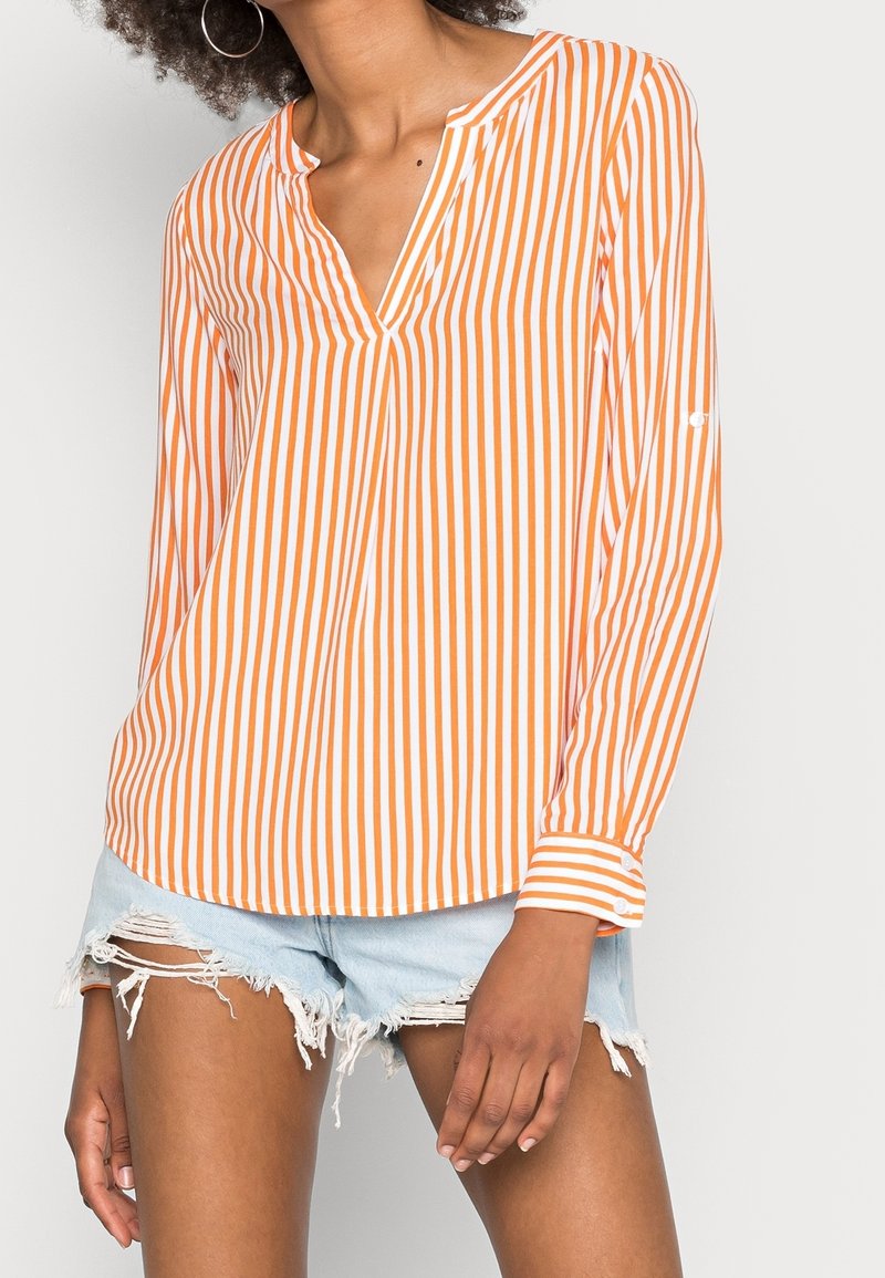 Camicia a maniche lunghe a righe arancioni e bianche con scollo a v e polsini abbottonati, indossata con pantaloni shorts di jeans strappati. Tessuto morbido e leggero.
