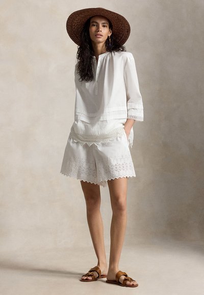 Γυναίκα που φορά καπέλο με φαρδύ γείσο σε καφέ χρώμα, λευκό πουκάμισο με μακριά μανίκια, λευκά σορτς από δαντέλα eyelet και καφέ σανδάλια, στέκεται με τα χέρια στις τσέπες.