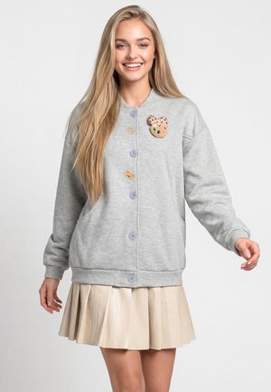 Ung kvinde med langt blondt hår iført en grå knapcardigan med farverige patches og et beige plisseret skørt, smilende.