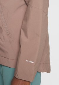 Manche de veste marron avec étiquette "DRYVENT" portée par-dessus un pantalon turquoise sur un fond clair uni.