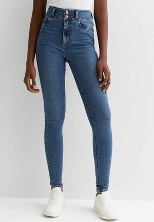 Jean en denim bleu coupe slim avec taille haute et fermeture à double bouton porté avec des baskets blanches et un haut blanc sur une personne debout.