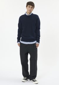Granatowy sweter z ribbami na mankietach i u dołu, noszony na jasnobłękitnej, w paski koszuli. Ciemne, luźne spodnie i czarne sneakersy dopełniają całość.