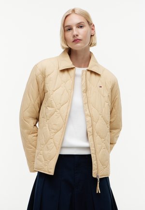 Blondynka w beżowej pikowanej kurtce zapinanej na zamek, na białej bluzce i granatowej plisowanej spódnicy, stojąca na jednolitym tle.