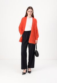 Lys oransje, fluffy cardigan over en hvit skjorte, kombinert med svarte stripete vide bukser og svarte spisse hæler, holder en svart veske.