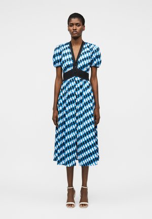 Γυναίκα που φοράει μεσοφόρι φόρεμα με μπλε, μαύρο και λευκό σχέδιο, με φουσκωτά μανίκια, μαύρη ζώνη στη μέση και λευκά σανδάλια με τακούνι, στέκεται μπροστά σε μονόχρωμο φόντο.