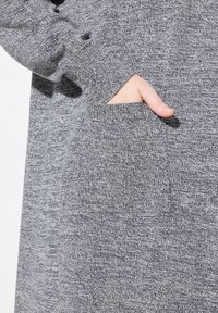 Grå texturstoff med en synlig ficka som visar en hand. Materialet verkar mjukt och stickat, med ett blandat mönster genom hela.