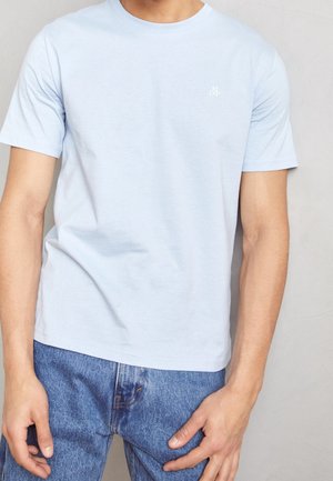 Personne portant un t-shirt à manches courtes bleu clair avec un petit logo blanc sur la poitrine et un jean en denim bleu sur un fond uni.