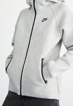 Jasnoszara bluza z kapturem Nike zapinana na zamek, z czarnym logo, czarnym suwakiem i kieszeniami na zamek, na osobie z rękami w kieszeniach.