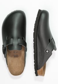 Sabots en cuir noir avec une bride et une boucle, une semelle intérieure en liège, une semelle extérieure texturée et une surface supérieure lisse. Logo de marque bien visible sur le côté.