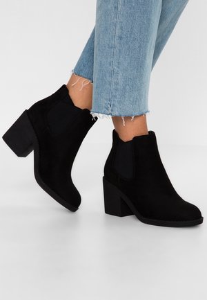 Personne portant un jean bleu clair à ourlet effiloché et des bottines en suède noir à talons épais sur fond blanc.