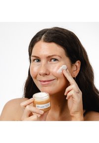 Hydratant facial dans un pot transparent avec une crème teintée orange. Une personne applique la crème sur sa joue, montrant une texture lisse et un éclat subtil.