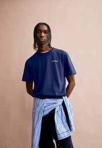 T-shirt en coton bleu marine avec un col rond, ornée d'un logo en texte blanc sur la poitrine. Chemise rayée bleu clair nouée autour de la taille.