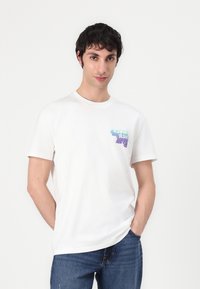 Hvid bomuld t-shirt med et farverigt grafisk print på venstre bryst. Korte ærmer, rund halsudskæring, afslappet pasform. Bæret med blå jeans.