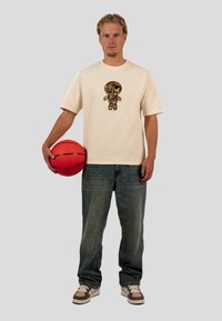 Cremfarbener Baumwoll-T-Shirt mit einem bunten grafischen Design, kombiniert mit blauen Jeans und braunen sowie weißen Sneakers, während er einen roten Basketball hält.