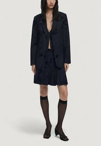 Mørk marineblå blazer med blomstret tekstur, kombineret med en matchende kort plisseret nederdel. Sorte halv-gennemsigtige knæstrømper og sandaler med åbne tæer.