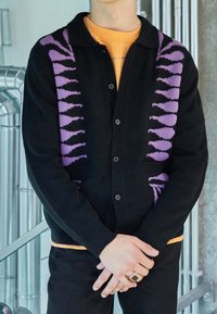 Svart cardigan med lila geometriskt mönster på axlarna, knäppt fram, och ribbade ärmslut. Bärs över en orange skjorta.