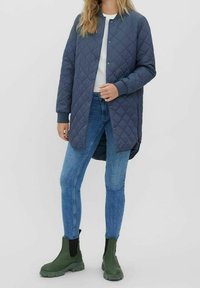 Quiltad lång jacka i mörkblått, med rund halsringning, tryckknappar och ribbad mudd, tillsammans med ljusblå skinny jeans och gröna ankelstövlar.