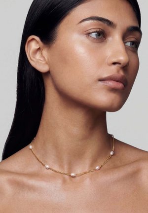 Nő sima bőrrel és hosszú sötét hajjal, aranyláncot visel, amelyen egyenletesen elhelyezkedő kis fehér gyöngyök vannak, oldalra néz.