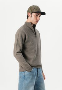 Bruin gebreid sweater met een hoge kraag, gecombineerd met een geruite pet. De outfit heeft een ontspannen pasvorm en geribbelde manchetten. Lichtblauwe jeans.