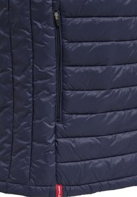 Zbliżenie na granatową pikowaną kurtkę z pionową zamykaną na zamek kieszenią i czerwoną metką marki w pobliżu dołu.