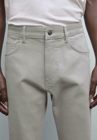 Pantaloni in cotone beige con taglio dritto, dotati di tasche frontali, chiusura con bottone e una sottile traspirabilità. Indossati con una blusa di colore chiaro.