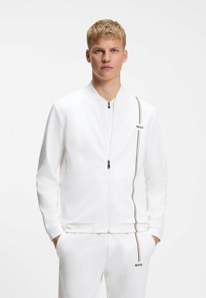Junger Mann trägt weiße Jacke mit Reißverschluss und passende Hose mit dünnen schwarzen und goldenen vertikalen Streifen und "BOSS"-Logos, Hände in den Taschen.