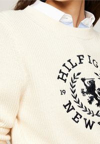Cream knit sweater with ribbed texture, featuring an embroidered black logo and laurel design at the front, worn over a white collared shirt.