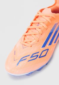 Scarpino da calcio sintetico arancione con accenti blu, caratterizzato da una superficie testurizzata, logo prominente e sistema di allacciatura tradizionale.