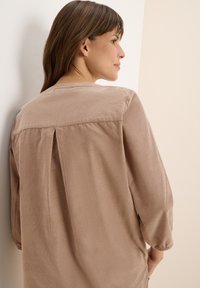 Femme aux cheveux bruns portant une chemise en velours côtelé beige à manches longues, tournée sur le côté contre un mur de couleur claire.