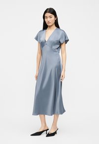 Γυναίκα που φοράει ανοιχτό γαλάζιο μεταξωτό midi φόρεμα με κοντά μανίκια και βαθύ V-λαιμόκοψη, συνδυασμένο με μαύρα μυτερά mule με τακούνι.