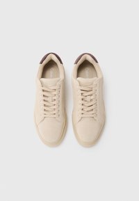 Beżowe zamszowe sneakersy z okrągłym noskiem, płaskimi sznurówkami i bordowym akcentem na pięcie. Charakteryzują się teksturowaną powierzchnią i logo wewnątrz.