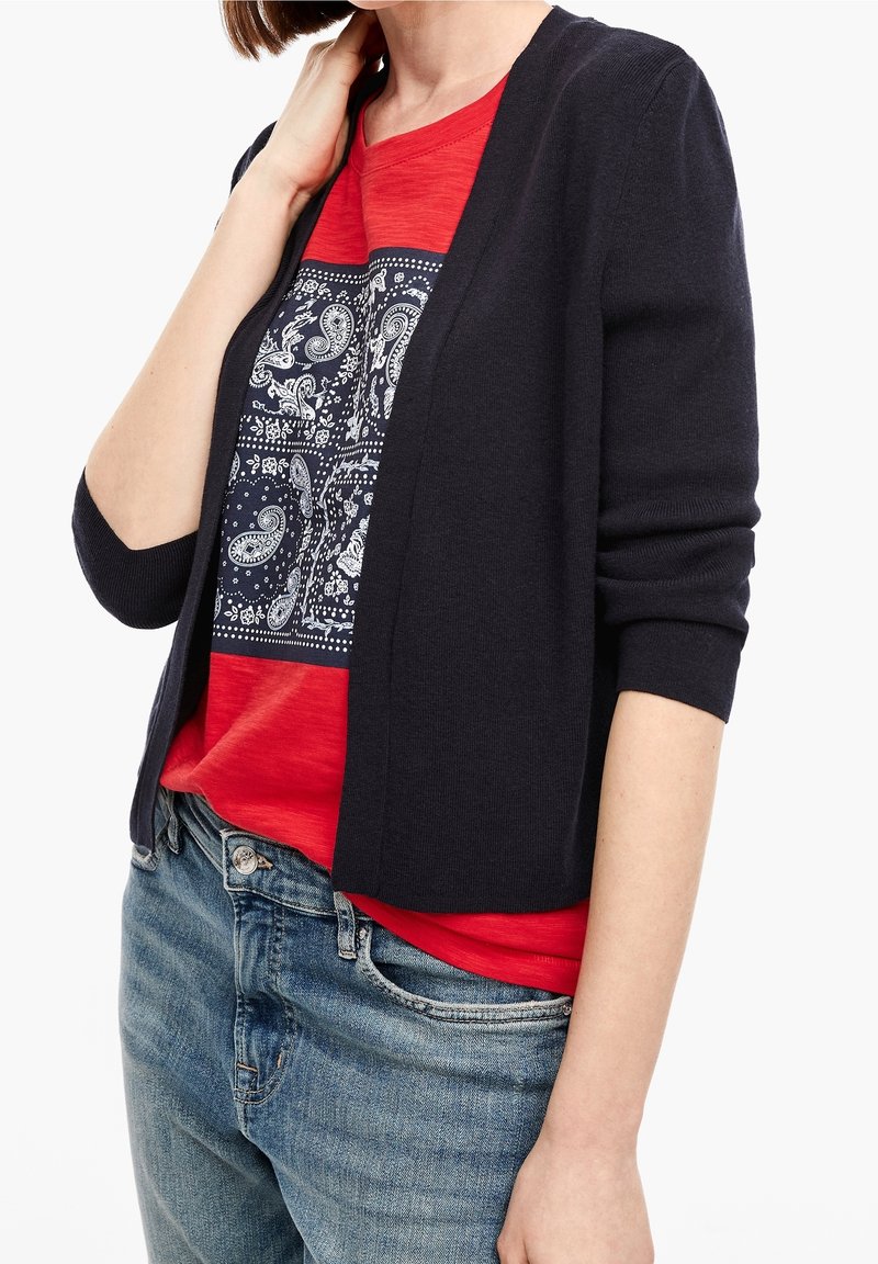 Femme portant un cardigan sombre ouvert sur une chemise rouge imprimée paisley avec un jean bleu, main reposant sur le cou, torse visible.