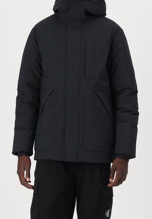 Veste matelassée noire avec capuche, fermeture éclair devant, deux poches devant et une poche poitrine sur une personne portant un pantalon noir.