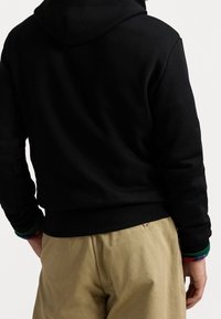 Schwarzer Baumwoll-Hoodie mit einer enganliegenden Säume, ausgestattet mit einer Kapuze und strukturierten Bündchen. Kombiniert mit hellbraunen, locker sitzenden Hosen.